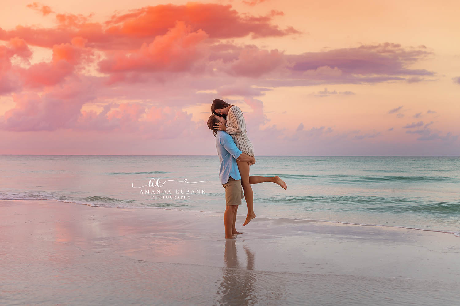 Seaside engagement photographer captures couple lifted in joyful embrace at pink sunset on the beach