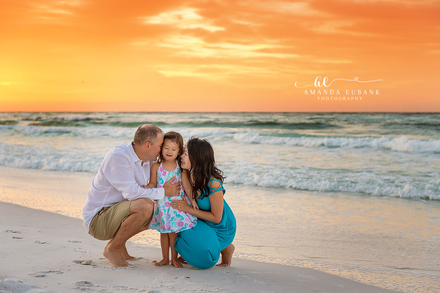 Seaside family photographer captures parents kissing toddler at water edge at vibrant orange sunset