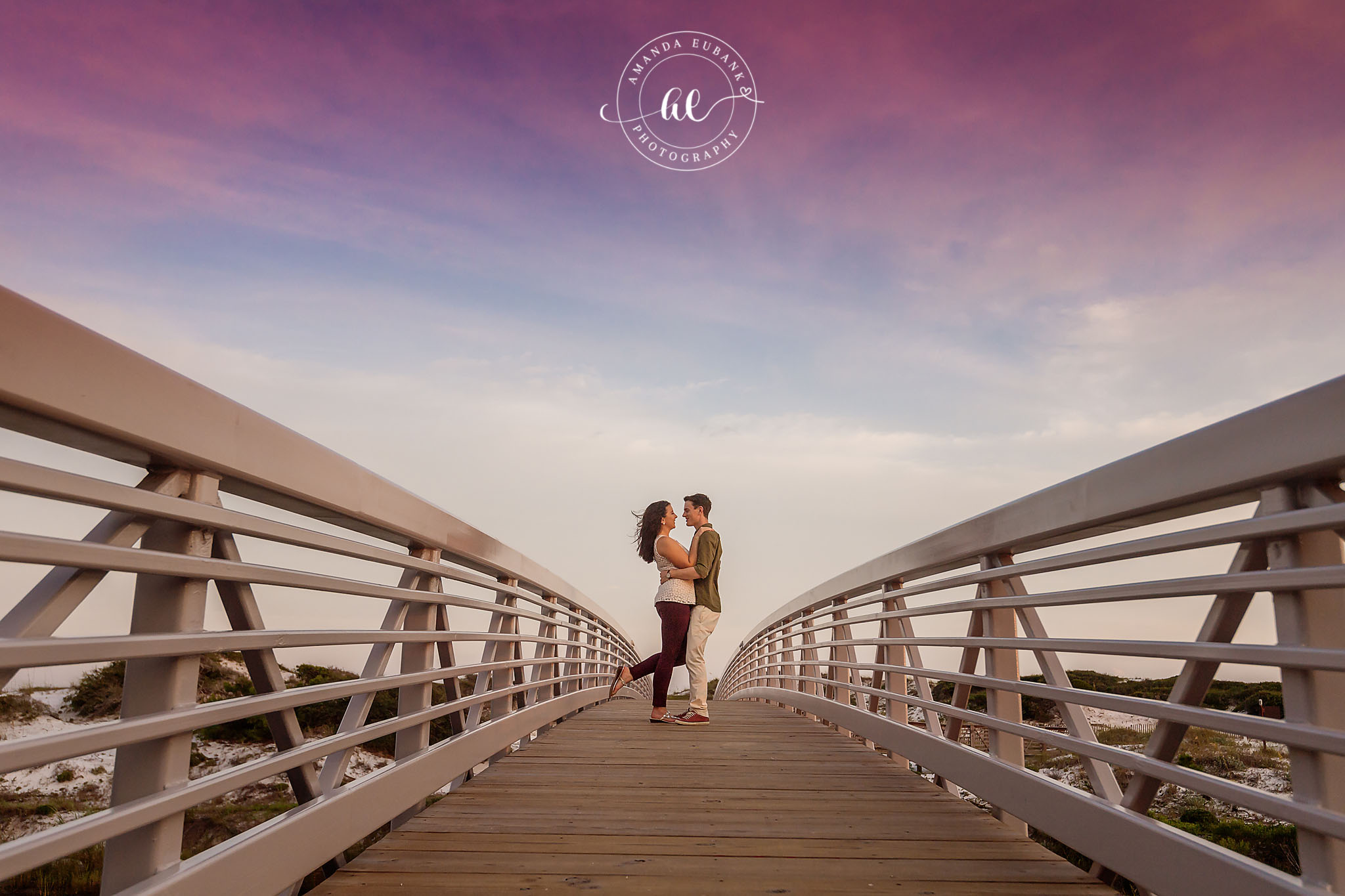 Seaside engagement photographer captures couple kissing on white pedestrian bridge at sunset