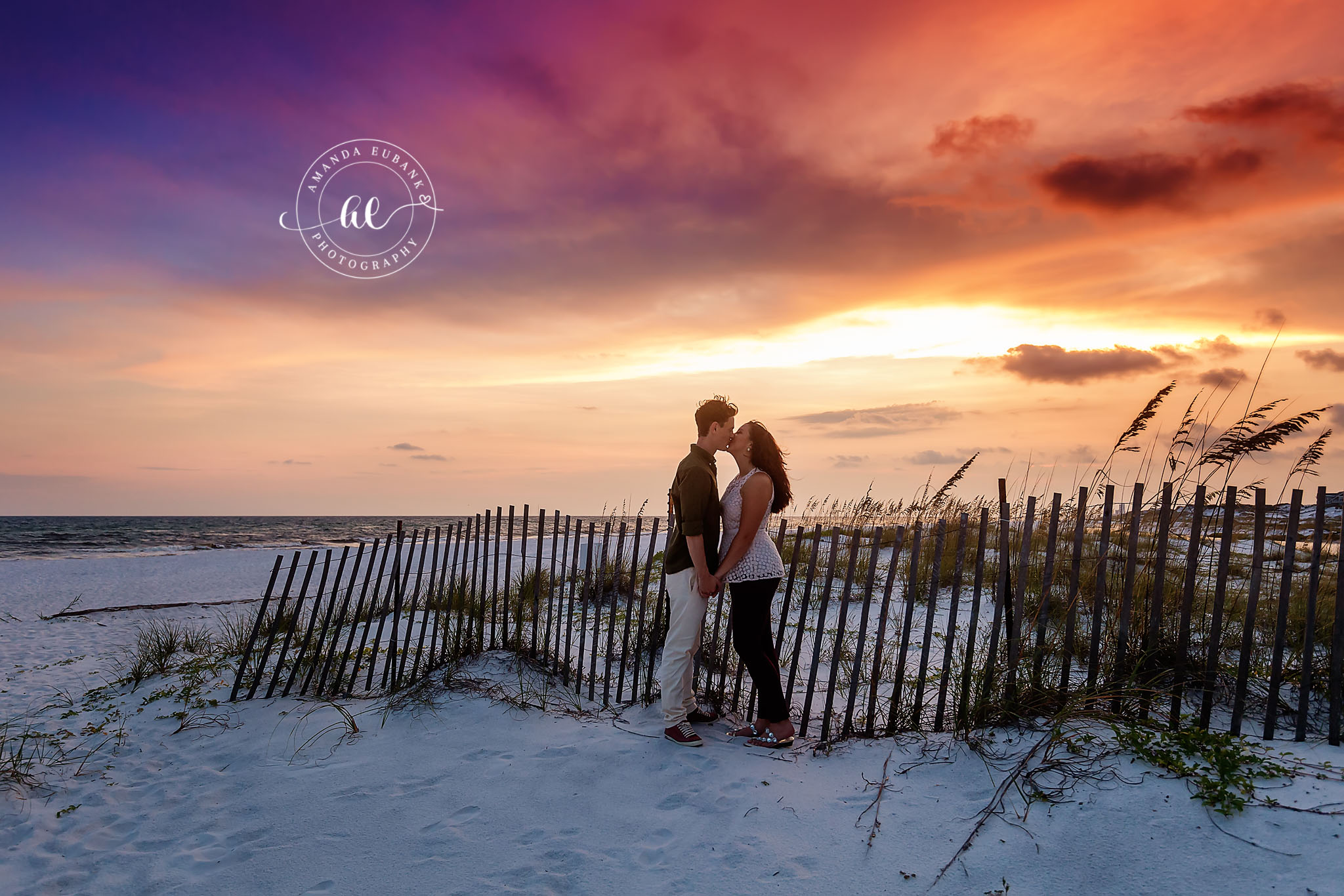 Seaside engagement photographer captures couple kissing by beach fence at dramatic sunset