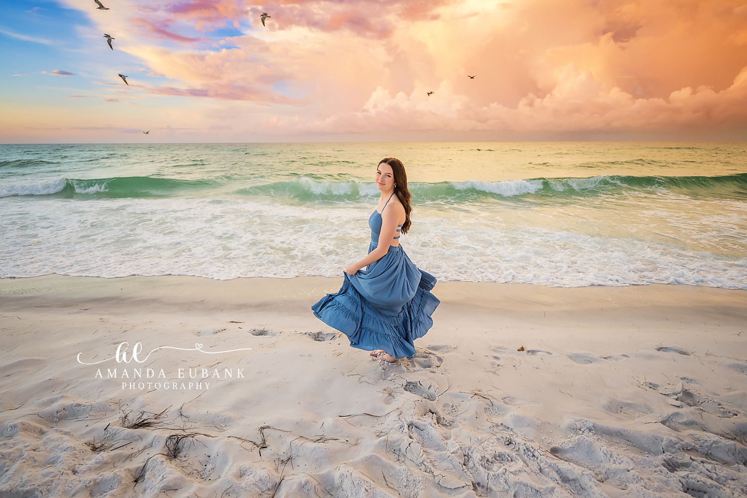 Seaside senior photographer captures brunette in flowing blue gown twirling on beach at sunset with seagulls