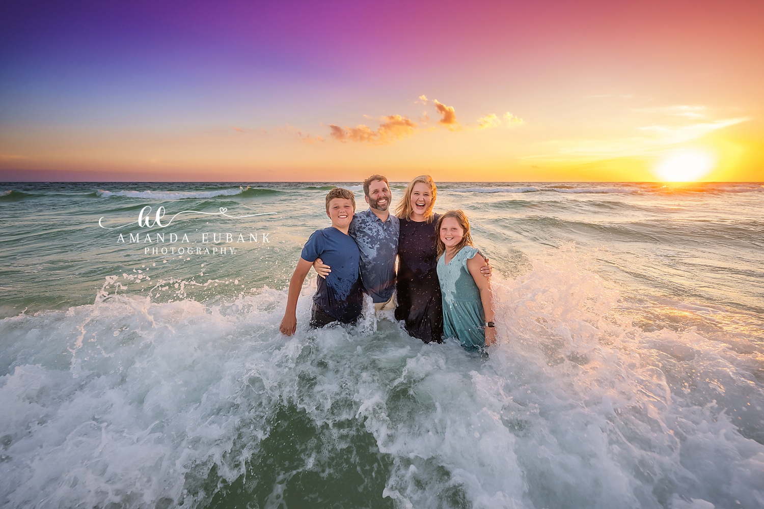 Seaside family photographer captures family of four in waist deep waves at vibrant sunset