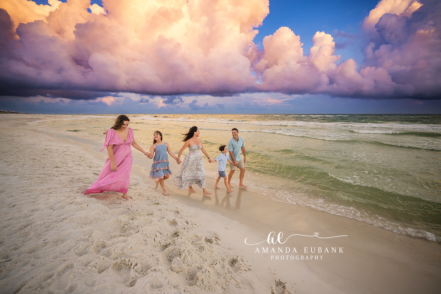 Seaside family photographer captures family of five walking shoreline under dramatic pink clouds