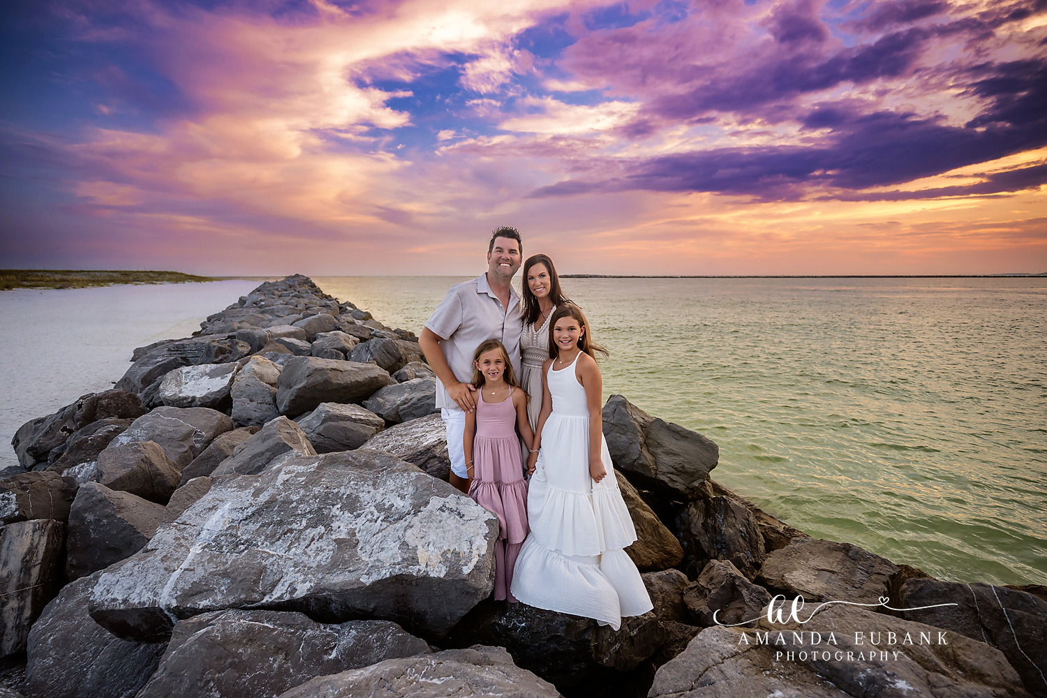 Seaside family photographer captures family of four on rocky jetty at vibrant sunset