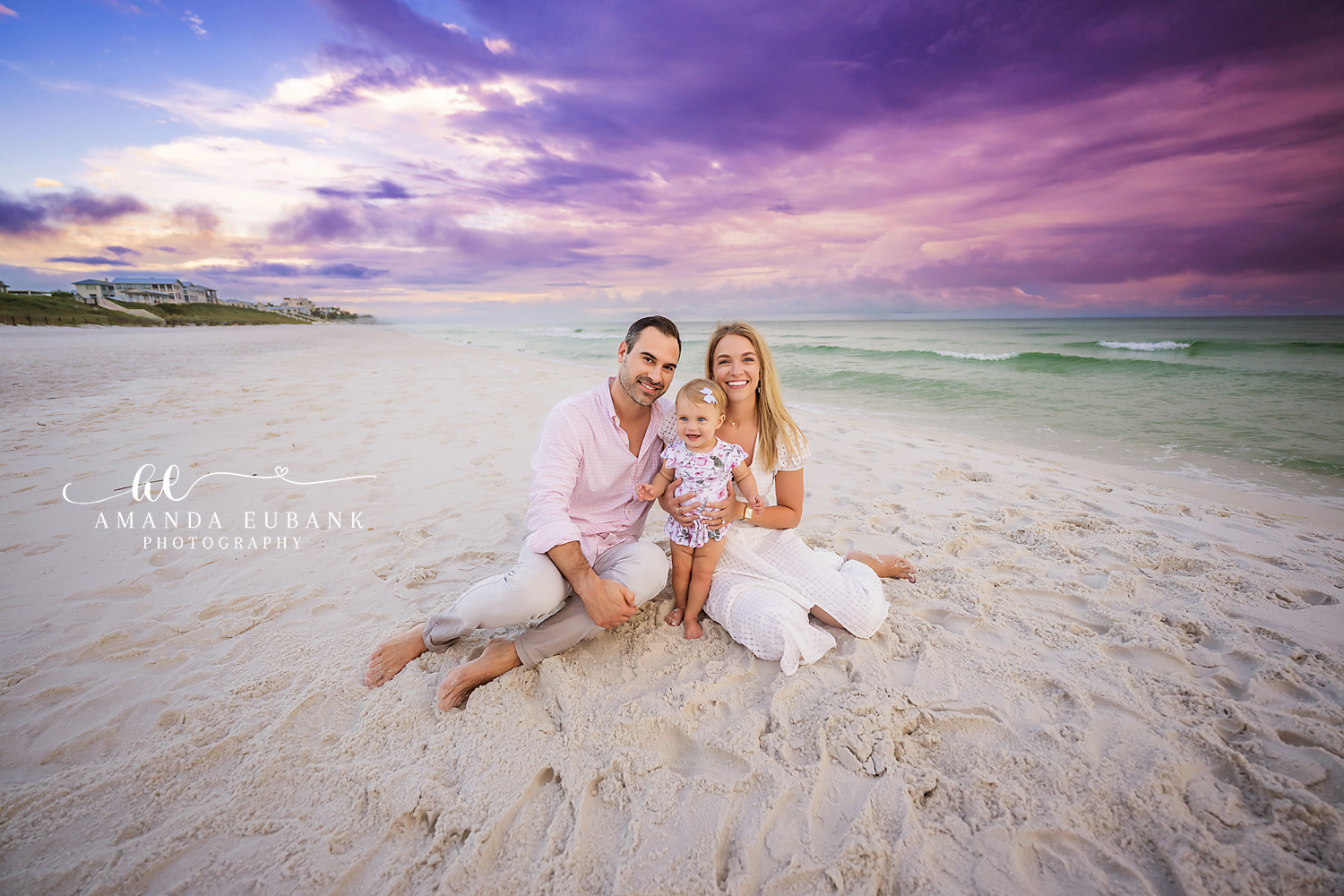 Seaside family photographer captures family of three with baby on beach at vibrant purple sunset