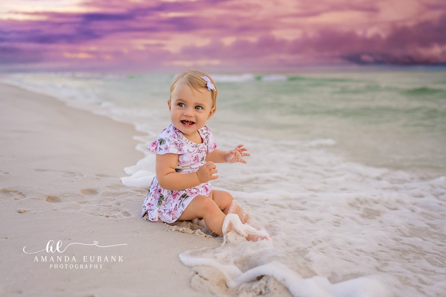 Seaside baby photographer captures baby girl in floral romper sitting in surf at purple sunset