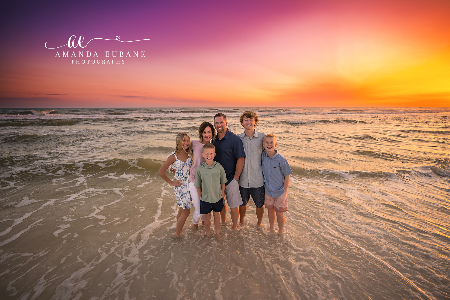 Seaside family photographer captures family of six at shoreline at vibrant orange and purple sunset
