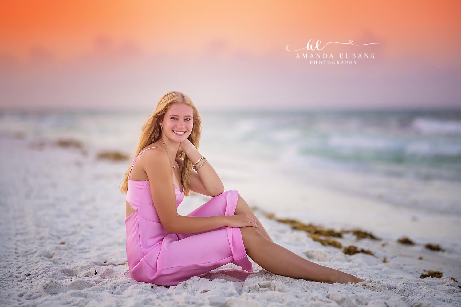 Seaside senior photographer captures senior in pink dress seated on the sand at peach sunset