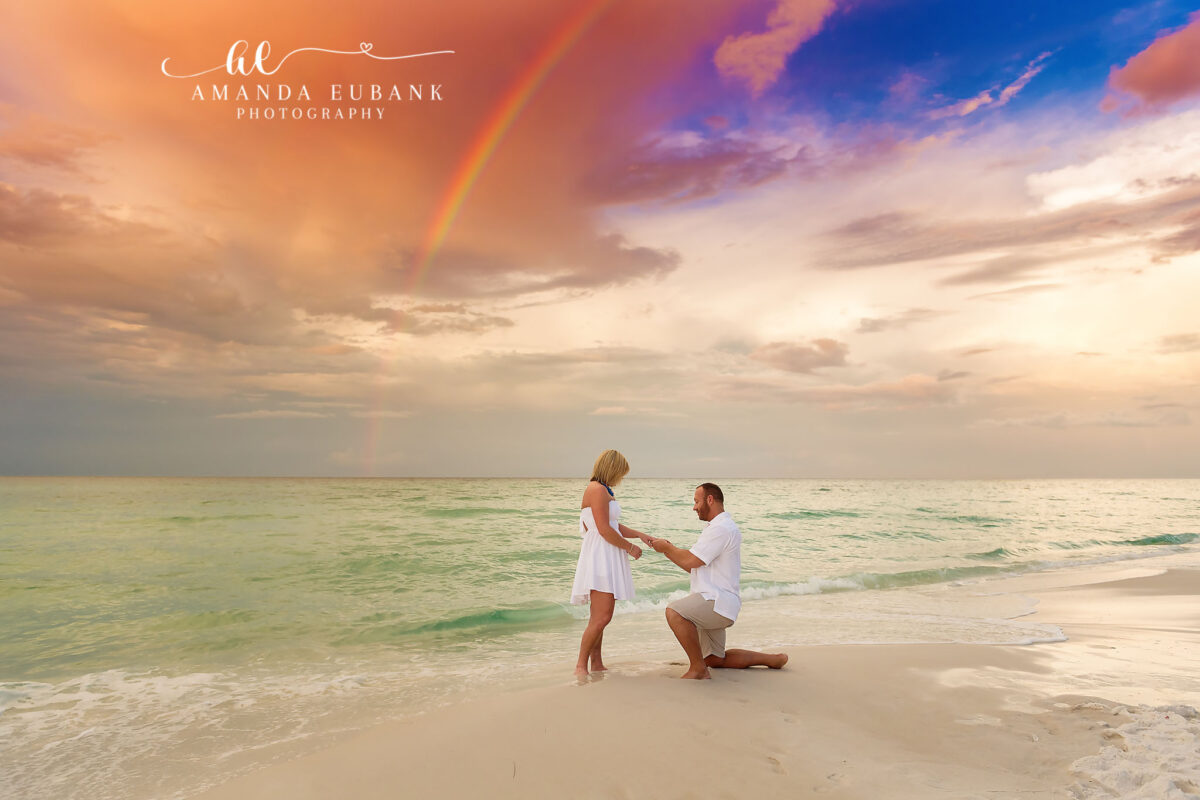 Man on one knee proposing to woman on the beach in Seaside Florida with a beautiful rainbow and dramatic golden sunset sky overhead