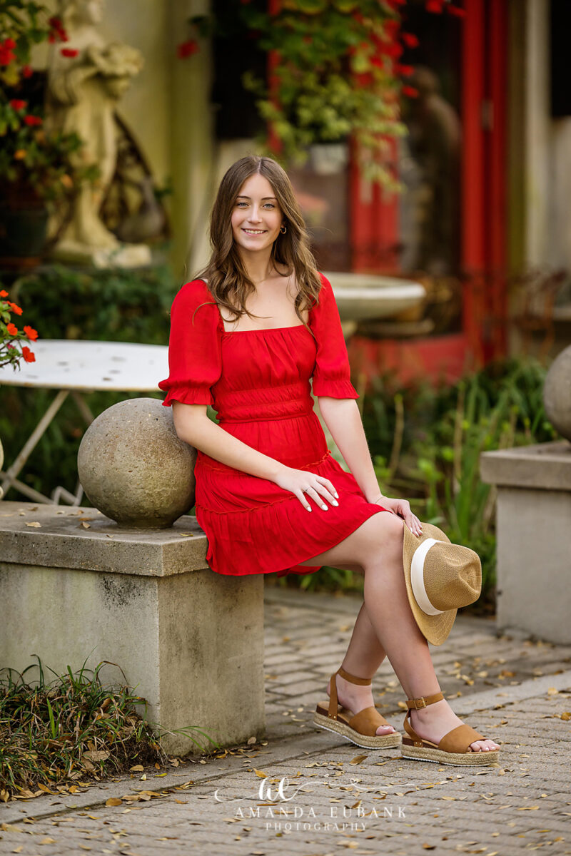 Senior girl in a red dress with dark brown hair sitting on a stone plinth in a colorful garden courtyard with a statue in Seaside Florida