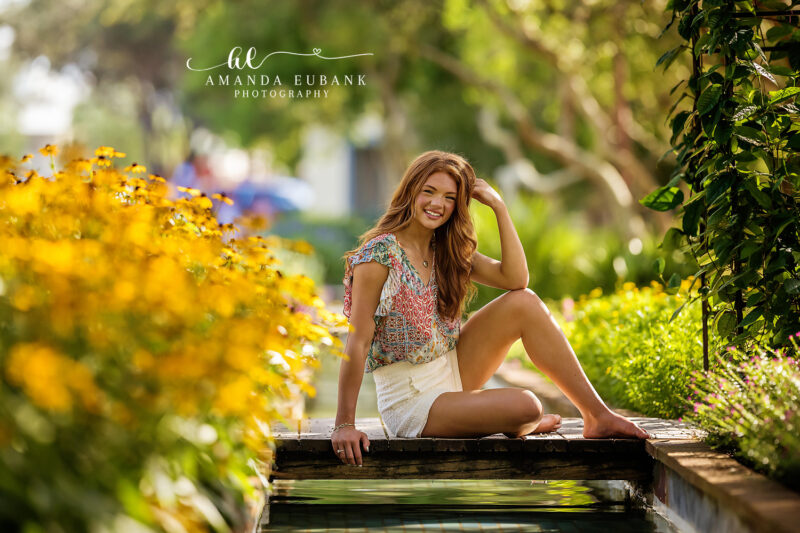 Senior girl in a blue dress sitting in the surf of the Gulf of Mexico at a warm golden and orange sunset in Seaside Florida