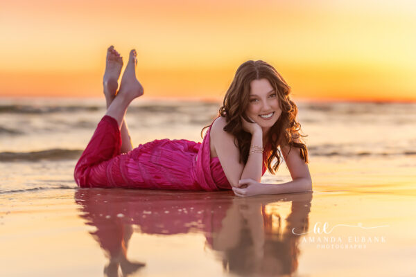 Senior girl with dark wavy hair in a blue maxi dress at the beach at a blazing golden and orange sunset sky in Seaside Florida